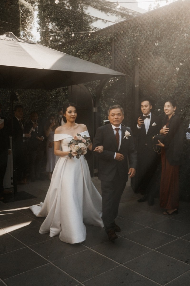 A bride walks through a garden strung with lights on her father's arm, guests standing either side