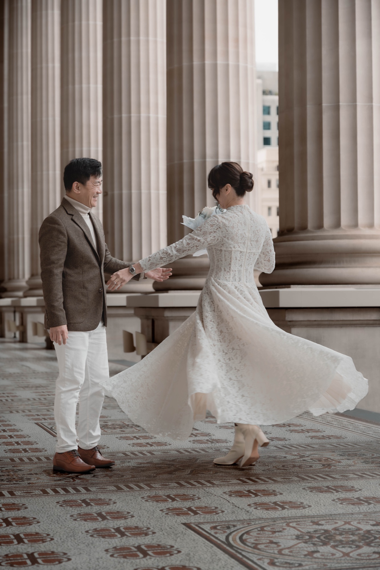 A bride twirls her lace dress beside her partner in the stone colonnade of Parliament House, Melbourne