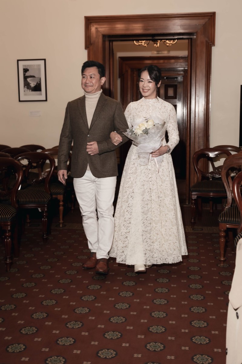 The couple walk in together, arm in arm, bouquet in hand