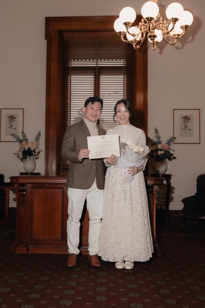 Newlyweds hold their marriage certificate and bouquet, beaming