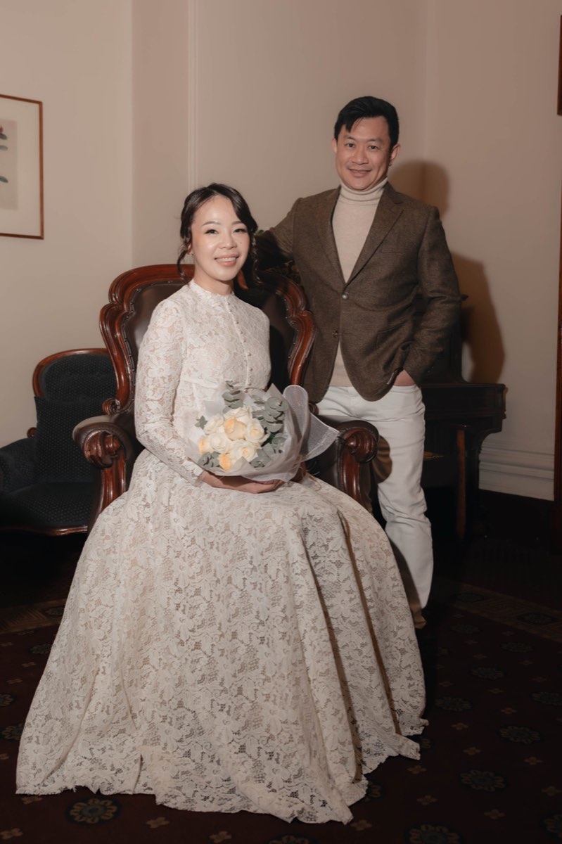 The bride seated in an antique armchair with her bouquet, the groom at her shoulder