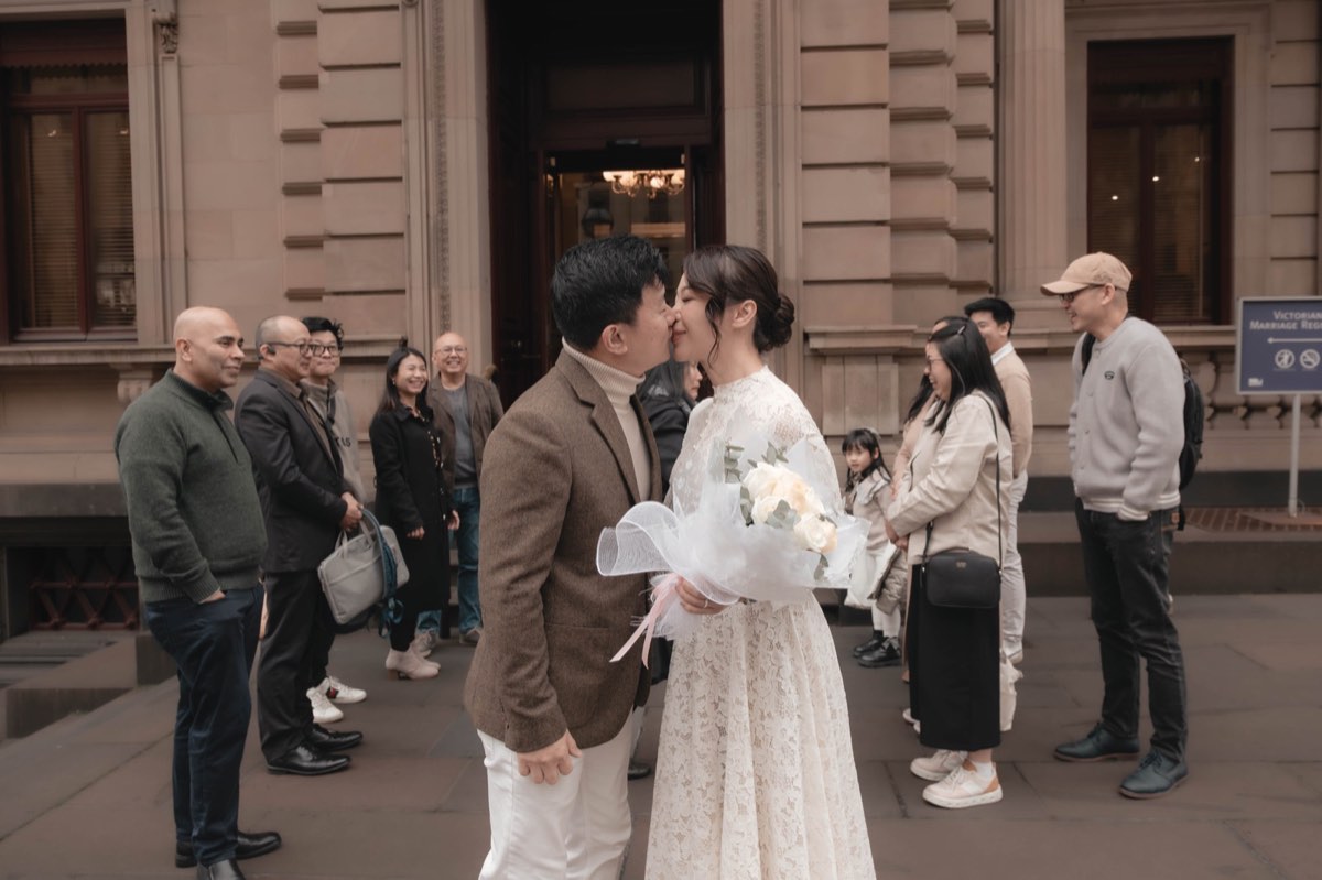The couple kiss on the footpath surrounded by their guests